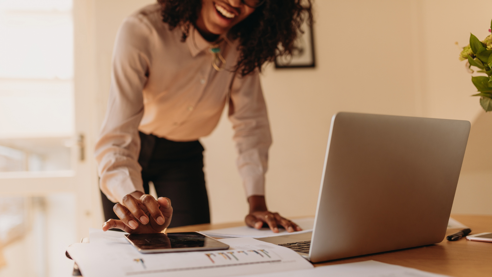 A smiling Black woman in a white blouse leans over a desk, tapping on a smartphone beside a laptop and printed charts in a bright home office setting