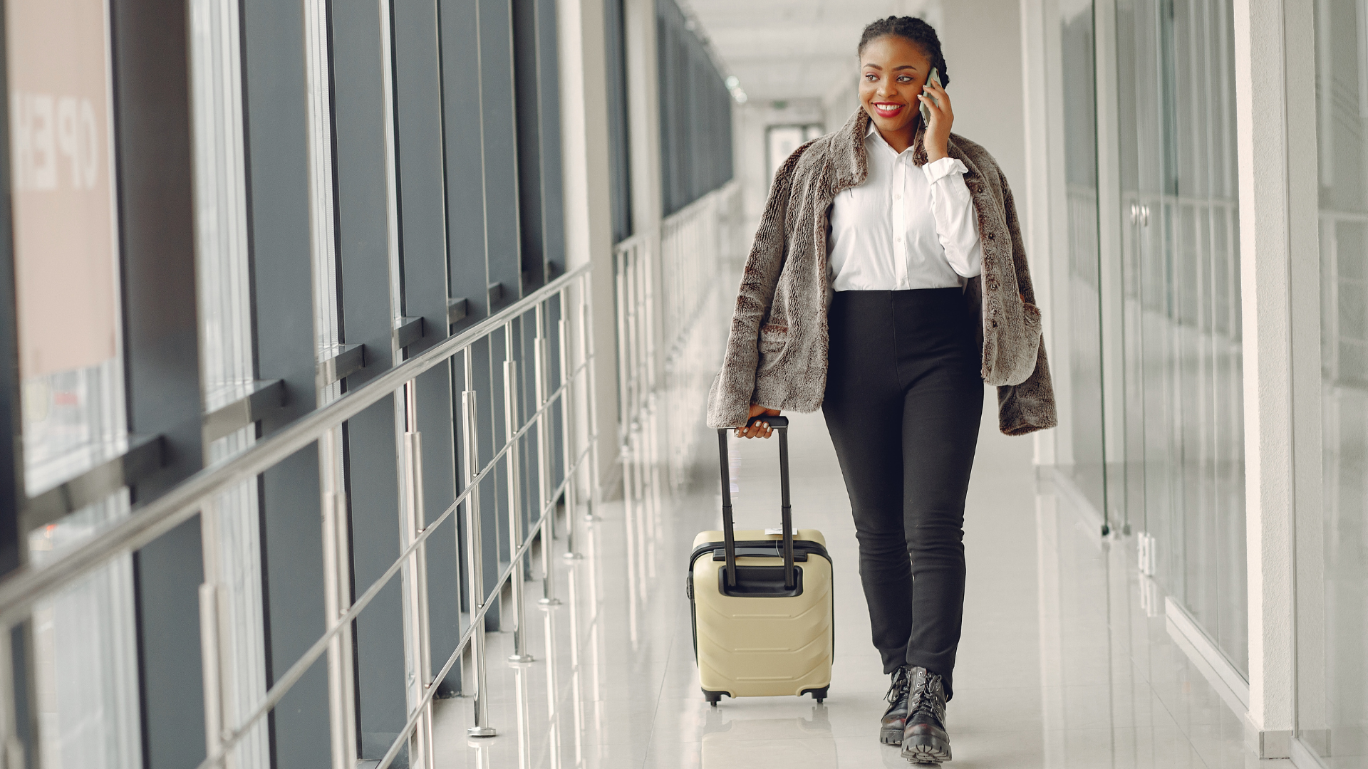 A smiling young Black woman walks through an airport corridor pulling a beige carry-on suitcase while talking on her phone, dressed in a white shirt, black pants, and a grey fur coat